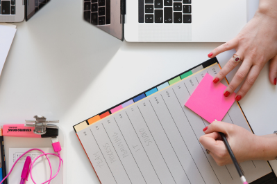 Overhead view of an event planner's hands writing on a sticky note and organizing a detailed project schedule in a planner on their desk.